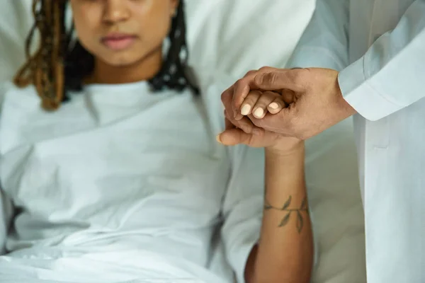 stock image cropped, doctor holding hand of african american woman in hospital gown, private ward, miscarriage