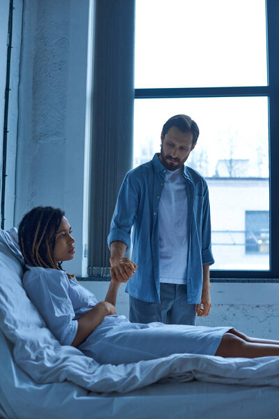 Концепция выкидыша, грустный мужчина держит за руку африканскую американскую жену, горе, больничную койку в частном отделении