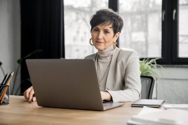 Modern ofis alanında dizüstü bilgisayarda çalışan orta yaşlı şık bir iş kadını.