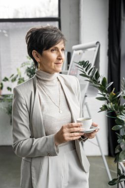 Kendine güvenen ve deneyimli orta yaşlı girişimci kahve fincanı ile modern ofise bakıyor.