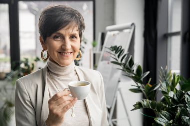 Kahve fincanı taşıyan neşeli orta yaşlı iş kadını ofisteki kameraya bakıyor, profesyonel vesikalık fotoğrafı.