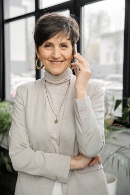 Yetişkin girişimci akıllı telefondan konuşuyor, kameraya gülümsüyor, profesyonel vesikalık çekiyor.