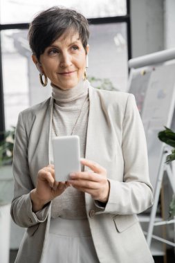 Kulaklıklı orta yaşlı bir iş kadını elinde akıllı bir telefonla ofisinden uzağa bakıyor.