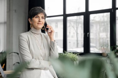 Kendine güvenen, orta yaşlı girişimci modern ofiste cep telefonuyla konuşuyor, başka tarafa bakıyor.