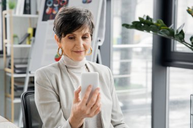 Mutlu olgun bir kadın, yönetici yönetici, modern çalışma ortamında cep telefonuna bakıyor.