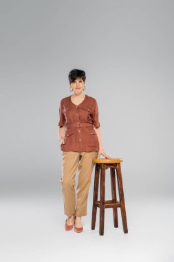 autumn fashion, middle aged woman in trendy clothes near high stool on grey, full length clipart