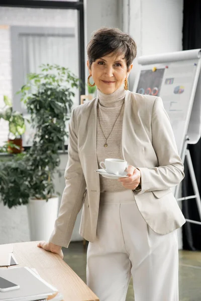happy middle aged businesswoman with coffee cup near smartphone and documents in office, portrait
