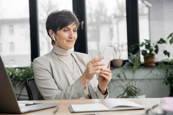 stock image pleased mature businesswoman in earbud browsing internet on smartphone near laptop in office