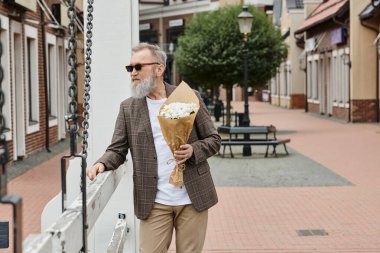 Sakallı, güneş gözlüklü, elinde çiçek buketi olan son sınıf öğrencisi. Şehir arkaplanı, şık kıyafetler.