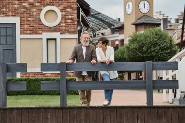 happy elderly couple, woman looking at man, standing near fence, urban backdrop, aging population clipart