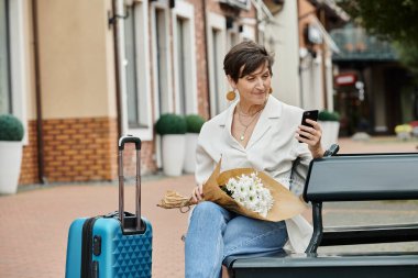 Kısa saçlı yaşlı bir kadın akıllı telefon kullanıyor, elinde buket tutuyor, bavulun yanında bankta oturuyor.