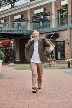 Mutlu ve sakallı yaşlı bir adam akıllı telefondan konuşuyor, alışveriş torbaları tutuyor, mağazanın yakınında yürüyor.