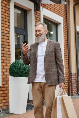 Sakallı neşeli yaşlı adam akıllı telefon kullanıyor, alışveriş torbaları tutuyor, prizin yanında yürüyor.