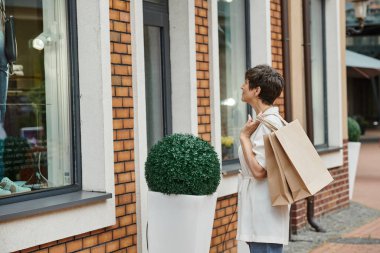 Kısa saçlı, alışveriş torbası tutan ve açık hava alışveriş merkezinin vitrinine bakan son sınıf öğrencisi kadın.