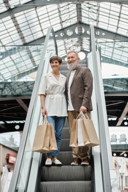 Neşeli yaşlı adam ve kadın yürüyen merdivende duruyor, alışveriş torbaları, alışveriş merkezindeki kameraya bakıyor