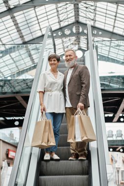 cheerful senior man and woman standing on escalator, shopping bags, looking at camera in mall clipart