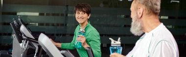 happy woman looking at elderly man, husband and wife in gym, holding sports bottles, banner clipart