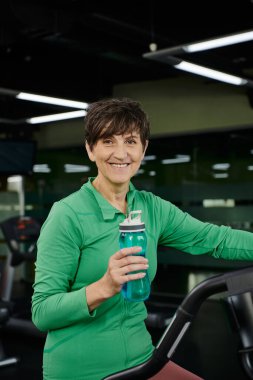 Mutlu yaşlı kadın elinde suyla spor şişesi tutuyor ve kameraya, spora, spor salonuna bakıyor, spor yapıyor.