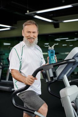 Yaşlı ve sakallı adam elinde su şişesiyle kameraya, spora, spor salonuna bakıp gülümsüyor.