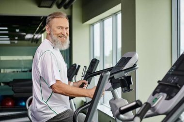 happy bearded man working out on exercise machine, elderly in gym, active senior, fitness and sport clipart