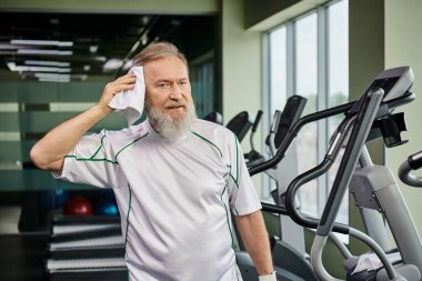 Spor salonunda spor yaptıktan sonra terini havluyla silen sakallı yaşlı adam kameraya ve spora bakıyor.