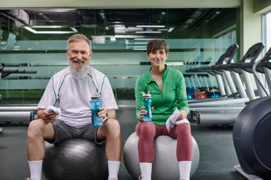 Sportif yaşlı çift, mutlu adam ve kadın fitness toplarının üzerinde oturmuş, ellerinde su şişeleriyle