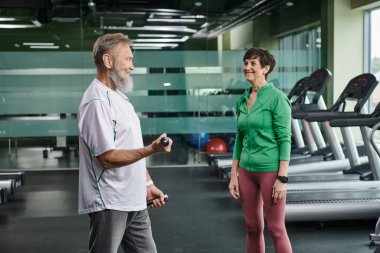 Sakallı kocaya bakan mutlu bir kadın spor salonunda dambıllarla çalışıyor, aktif yaşlılar, yaşam tarzı...