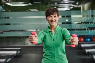 Kısa saçlı mutlu kadın, halterlerle çalışıyor, spor salonunda kameraya bakıyor, portre çiziyor.