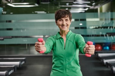 happy woman with short hair working out with dumbbells, looking at camera in gym, portrait clipart