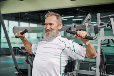 Sakallı, güçlü ve yaşlı bir adam spor makinesinde çalışıyor, atletik ve sağlıklı, spor salonu.