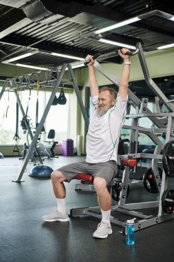 Sakallı, güçlü ve yaşlı bir adam spor makinesinde çalışıyor, atletik ve sağlıklı, uzun boylu.