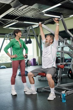 Sakallı, yaşlı ve güçlü bir adam egzersiz makinesinde çalışıyor. Yanında mutlu bir kadın, spor salonu, aktif.