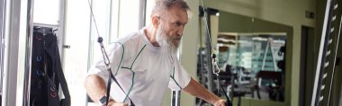 motivated elderly man with beard working out on exercise machine in gym, athlete, active, banner clipart