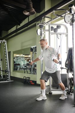 Sakallı yaşlı bir adam spor salonunda egzersiz makinesi, atlet ve motivasyon üzerinde çalışıyor.
