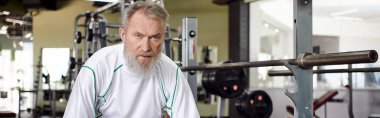tired elderly man with beard looking at camera after workout, exercise machine in gym, banner clipart