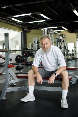 Egzersiz sonrası kameraya bakan sakallı, yorgun yaşlı adam spor salonunda egzersiz makinesinde oturuyor.
