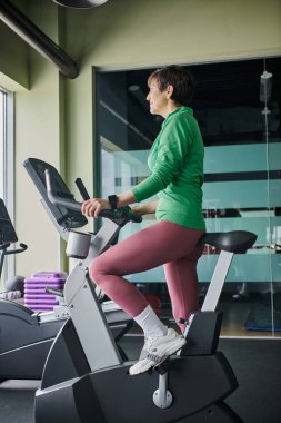 Kısa saçlı yaşlı bir kadın spor salonunda egzersiz bisikletine biniyor, son sınıf öğrencisi, motivasyonlu.