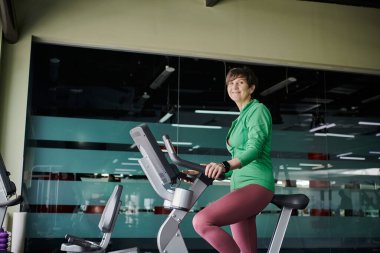 Kısa saçlı mutlu yaşlı kadın spor salonunda egzersiz bisikleti üzerinde çalışıyor, aktif, motivasyonlu.