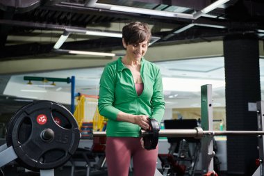 Yaşlı kadın gülümsüyor ve ağırlık plakası ile halterin yanında duruyor, spor salonunda çalışıyor, motivasyon