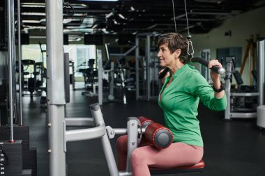 Sportif ve motive olmuş yaşlı kadın spor salonunda çalışıyor, olgun fitness, egzersiz makinesi, yan görüş