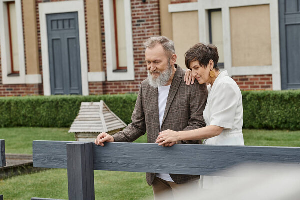 cheerful elderly couple, man and woman standing near fence next to house and looking away, romance