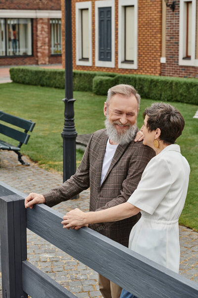 happy elderly couple, senior man and woman standing near fence next to house, looking at each other
