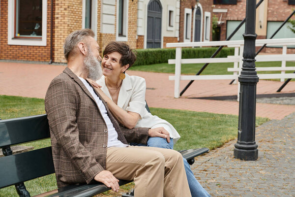 happy elderly woman looking at bearded man, romance, husband and wife sitting on bench, urban