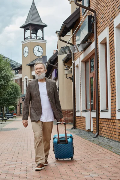Sakallı yaşlı adam şehir caddesinde bavulla yürüyor, şehir yaşam tarzı, seyahat, şık yaşlılar