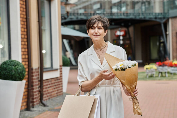 aging population, pleased senior woman holding shopping bags and bouquet near outlet, outdoors
