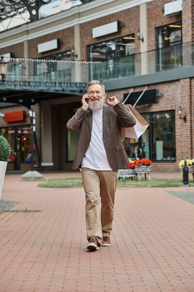 happy and elderly man with beard talking on smartphone, holding shopping bags, walking near outlet