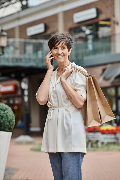 happy elderly woman talking on smartphone, holding shopping bags and standing near outdoor mall