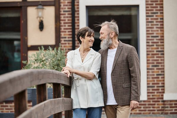 счастливый бородатый мужчина и женщина, глядя друг на друга, пожилая пара, стоя возле моста, романтика
