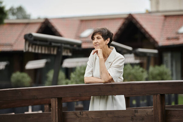 pensive senior woman standing on wooden bridge and looking away, elderly life, blurred background
