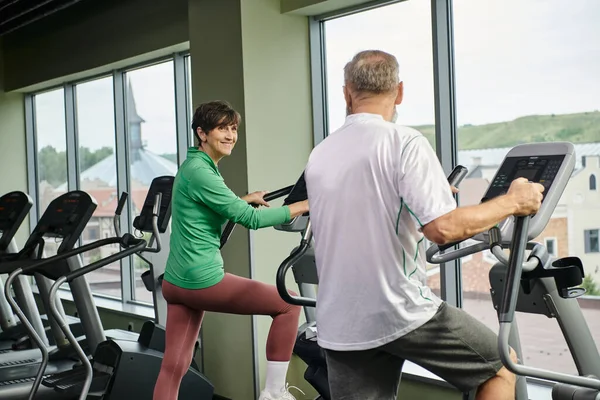 active seniors, happy woman looking at elderly man in gym, exercising together, senior couple